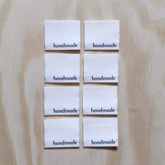 Eight premium woven garment labels arranged in 4 rows of 2 on a natural wood background. Labels are off-white, with 'handmade' woven in black on the bottom right-hand corner. Labels created by Stitch Collective.